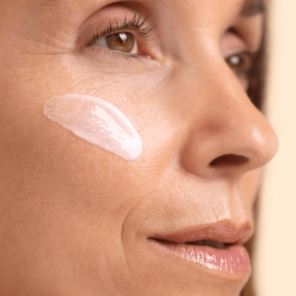 Close-up of a woman's face with a dollop of Ultra Sensitive Cream applied to her cheek against a soft beige background.