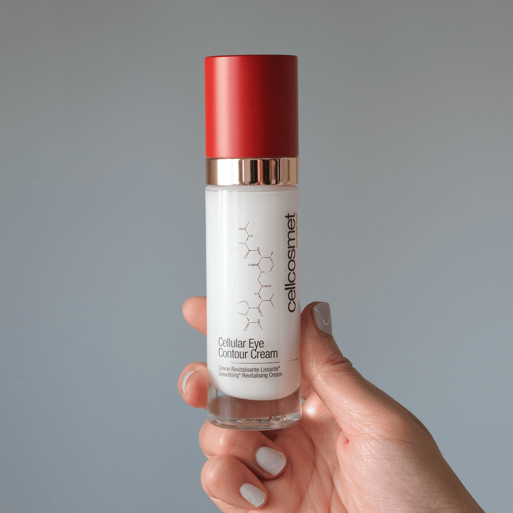 Hand holding a bottle of cellular eye contour cream with a red cap against a gray background