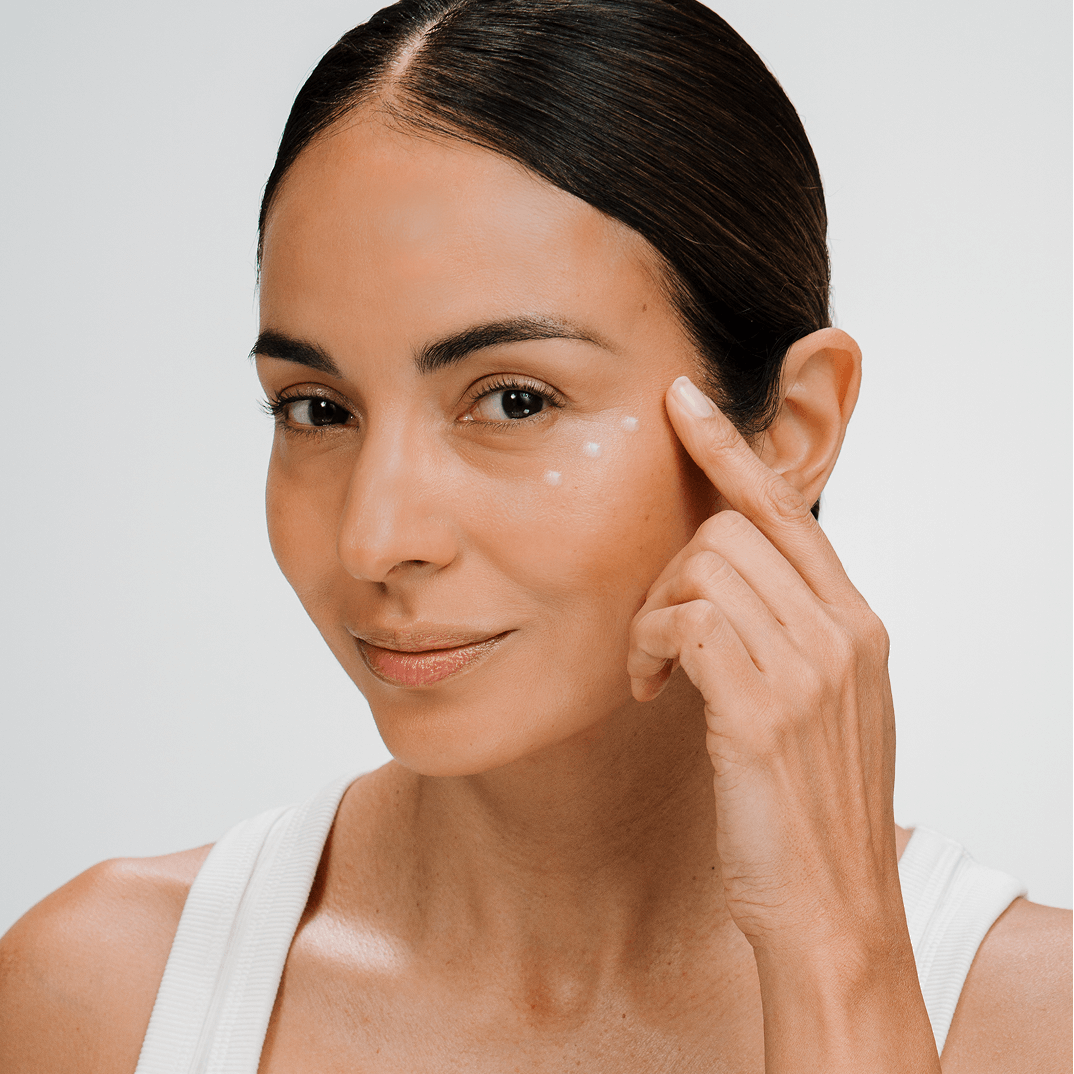 Woman applying cellular eye controur cream skincare product to her face against a neutral background